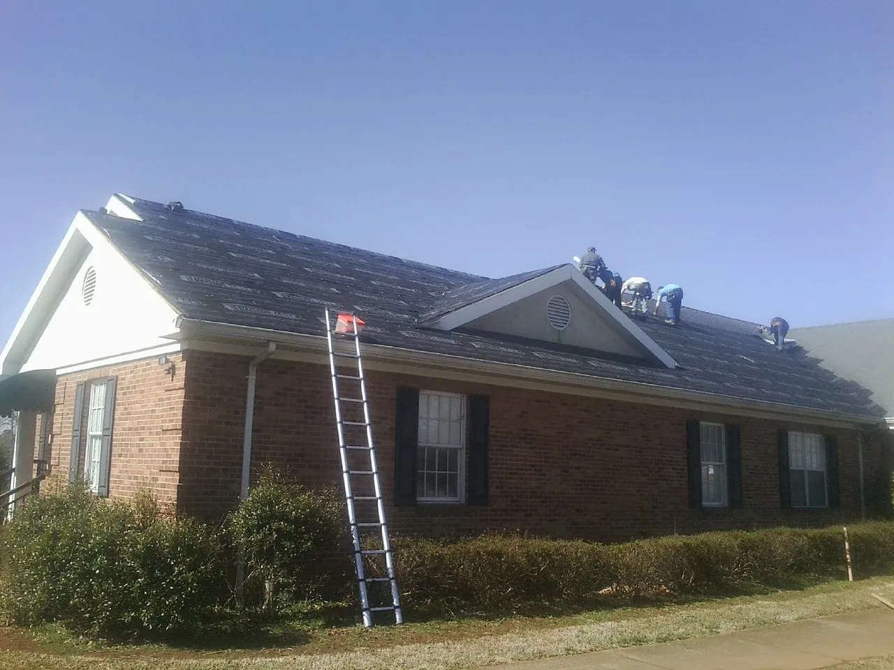 Crew tearing shingles off a brick ranch roof with a ladder set — Metal Roof Repair work by our crew in Longs