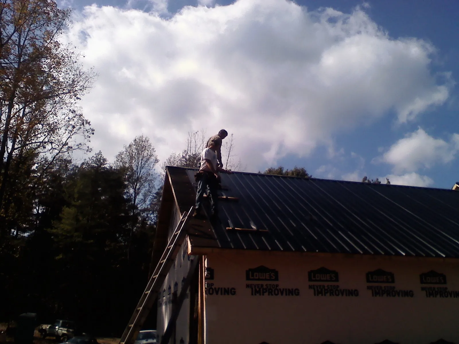 Roofer fastening dark metal panels high against a cloudy sky, from a Roof Inspection job in Longs